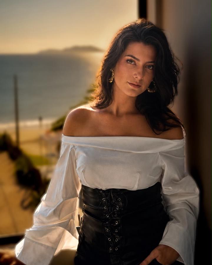 Kath Zaparoli, de blusa branca ombro a ombro e corpete preto, diante de uma janela com vista para o mar ao pôr do sol